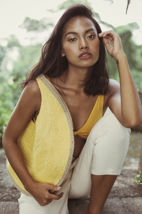 Woman wearing a yellow top and white pants sitting outdoors with greenery in the background