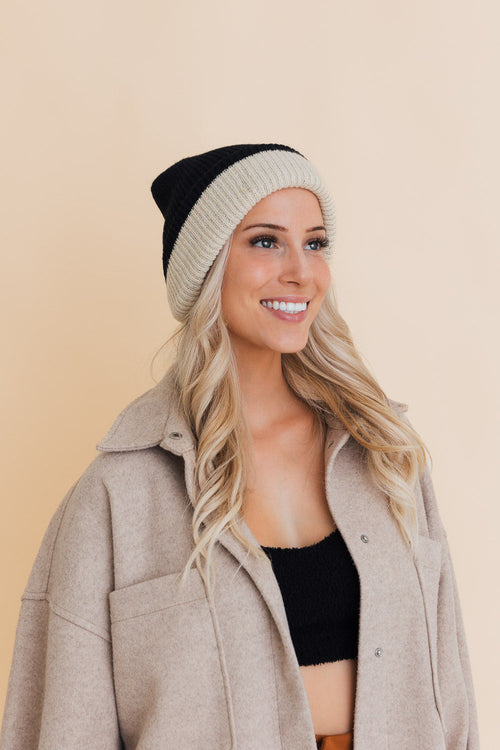 Woman wearing a beige coat, black top, and black and beige beanie against a beige background