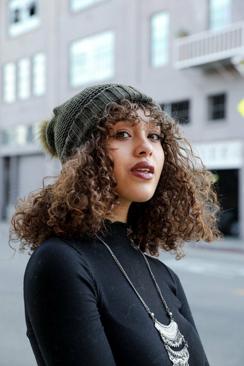 Woman wearing a green beanie and black sweater with curly hair, standing in an urban setting.