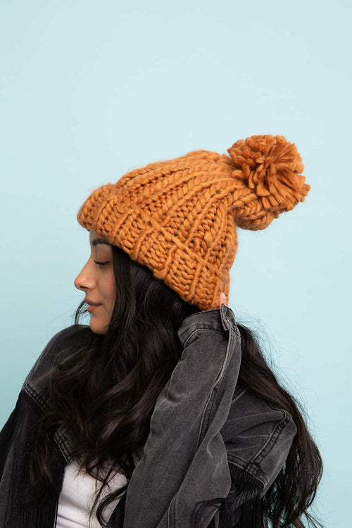 Person wearing a knitted mustard yellow beanie with a pom-pom against a light blue background