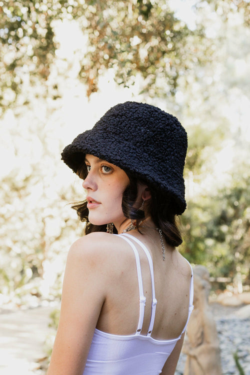 Woman wearing a black knit hat and white top in a natural setting with greenery.