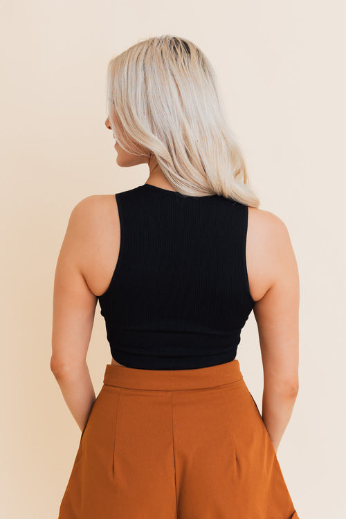 Woman wearing a black sleeveless top and brown pants against a beige background