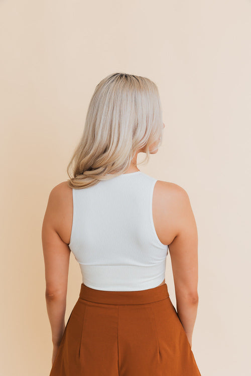 Person wearing a white sleeveless top and brown pants against a beige background