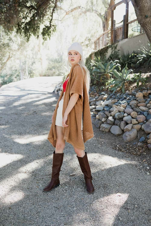 Woman in a brown poncho and knee-high boots standing on a gravel path with trees and rocks in the background.