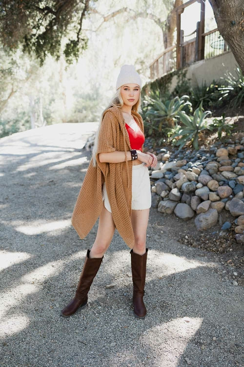 Woman in a casual outfit with a long cardigan, red top, white shorts, and brown boots standing outdoors.