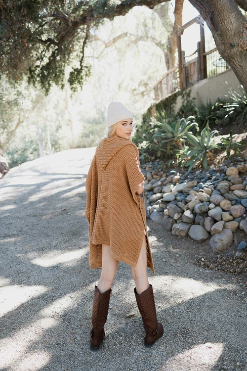Person wearing a brown coat and boots walking on a gravel path with trees and rocks in the background