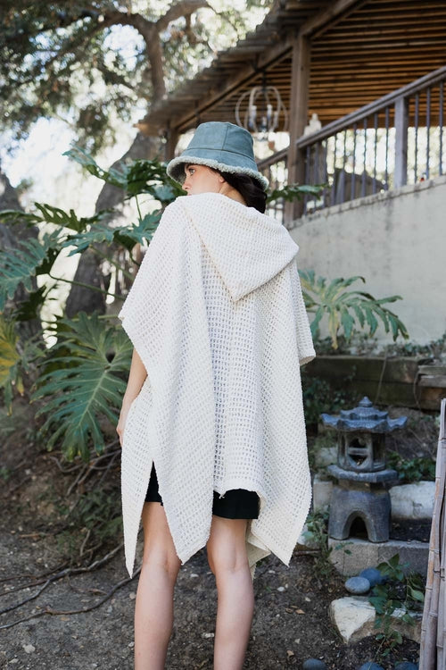 Person wearing a white knitted poncho and blue hat in an outdoor setting with plants and a building.