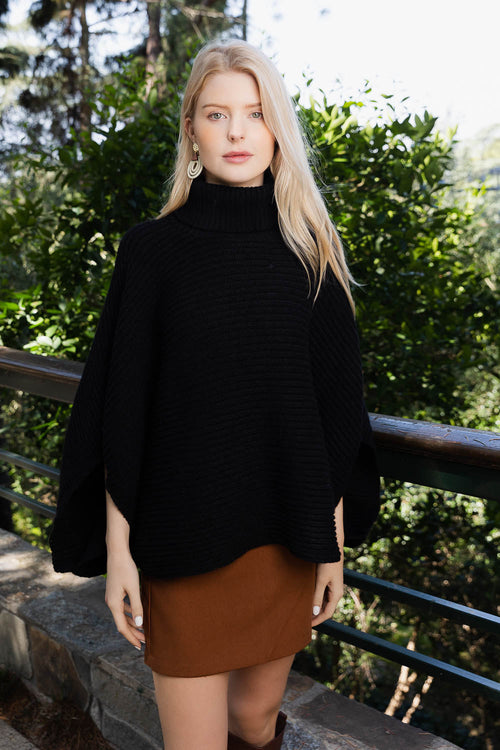 Woman wearing a black turtleneck sweater and brown skirt standing outdoors with greenery in the background