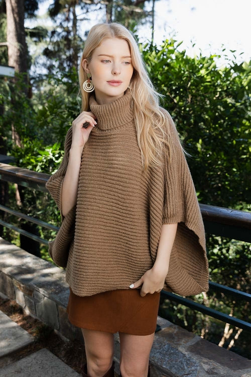 Woman wearing a brown poncho and skirt outdoors with greenery in the background