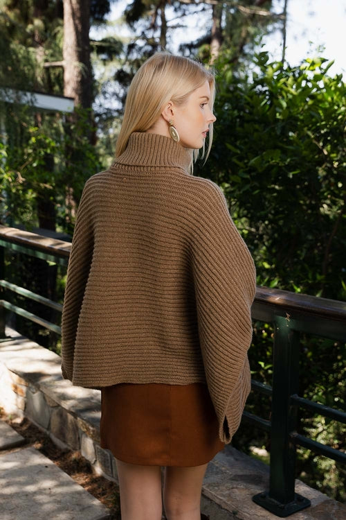 Woman wearing a brown knitted poncho standing outdoors with greenery in the background