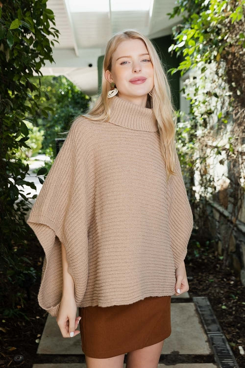Woman wearing a beige poncho outdoors with greenery in the background