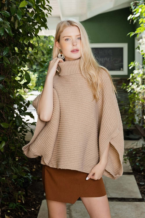 Woman wearing a beige poncho outdoors with greenery in the background