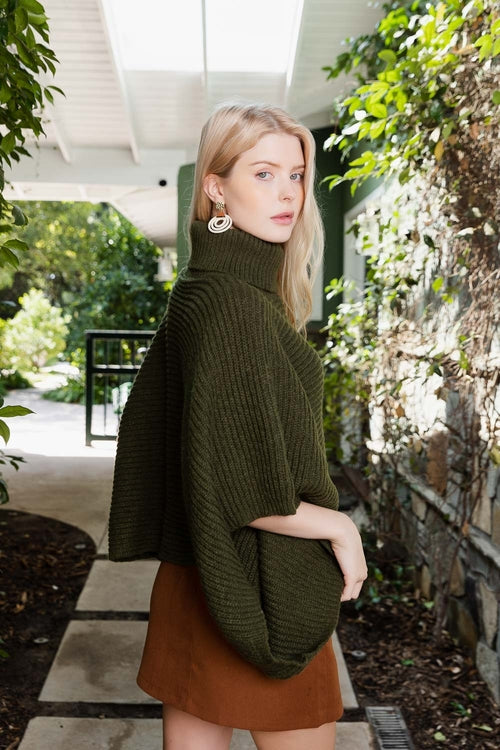 Woman wearing a green sweater and brown skirt standing outdoors with greenery around.
