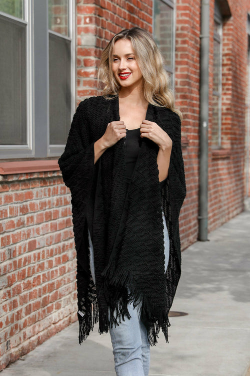 Woman wearing a black fringe cardigan against a brick wall.