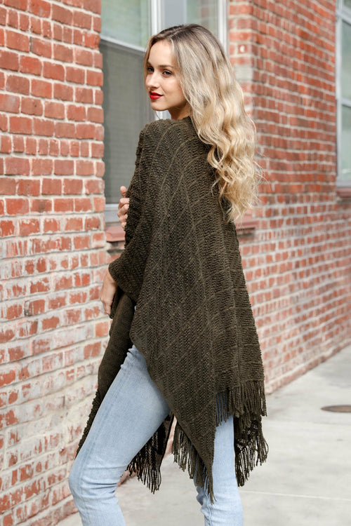 Woman wearing a brown fringed poncho against a brick wall.