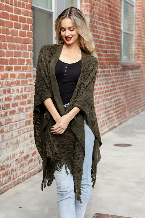Woman wearing a green poncho with fringe, black top, and light blue jeans against a brick wall.