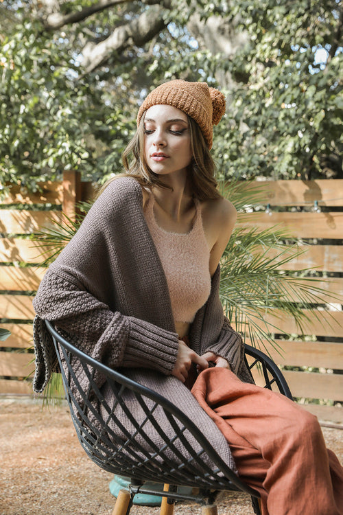 Woman sitting outdoors wearing a brown knit hat, sweater, and pants.