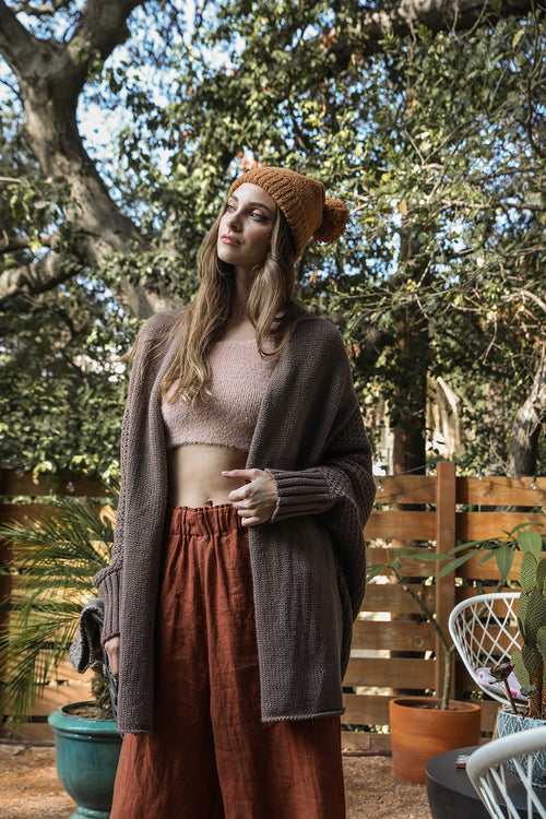 Woman wearing a brown beanie and cardigan in an outdoor setting with trees and plants.