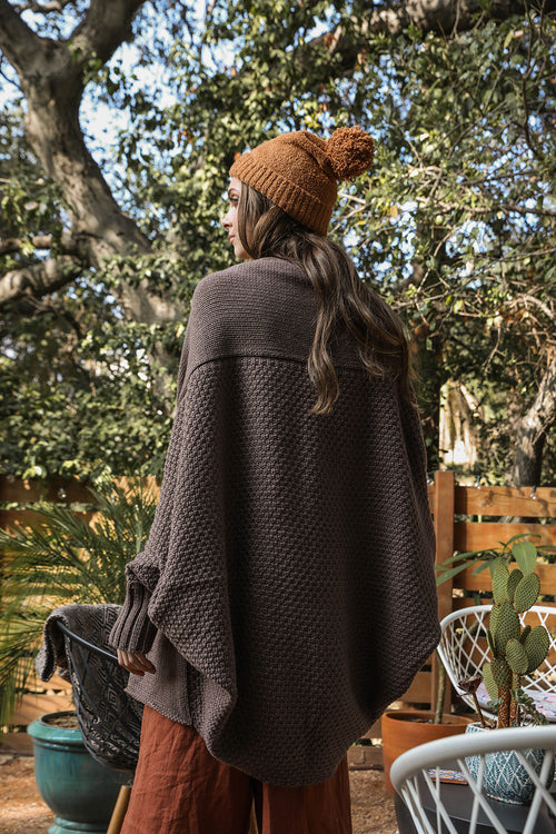 Person wearing a brown poncho and beanie standing outdoors with trees in the background