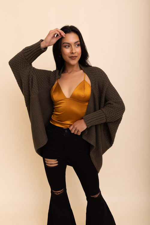 Woman wearing a mustard top and black pants with a brown cardigan against a beige background