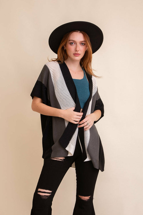 Woman wearing a black and gray patterned cardigan over a blue top and black pants, with a black hat on a beige background.