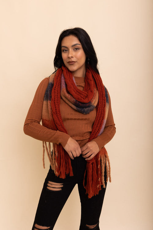 Woman wearing a brown sweater and black pants with a long, colorful scarf against a beige background