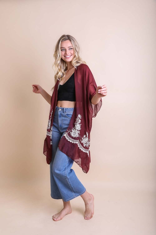 Woman wearing a burgundy kimono with white embroidery over a black top and blue jeans on a beige background