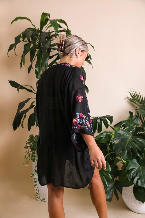 Person wearing a black floral kimono standing in front of green plants.