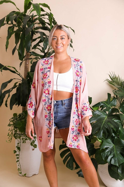 Woman wearing a floral kimono over a white top and denim shorts, standing in front of green plants.