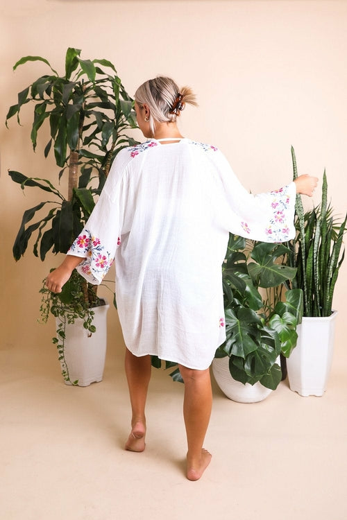 Woman wearing a white kimono with floral embroidery in a room with plants.