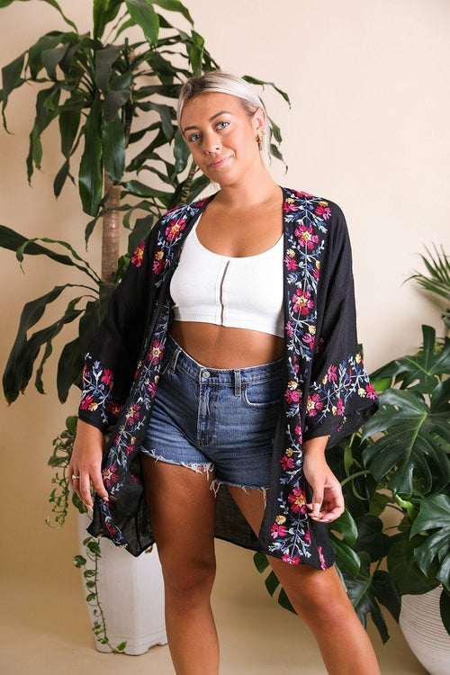 Woman wearing a floral kimono over a white top and denim shorts, standing in front of plants.