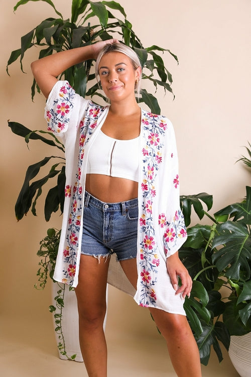 Woman wearing a floral kimono over a white top and denim shorts, standing in front of green plants.