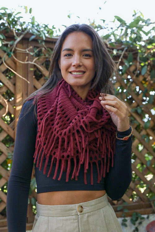 Woman wearing a burgundy knitted scarf in front of a wooden fence with greenery.