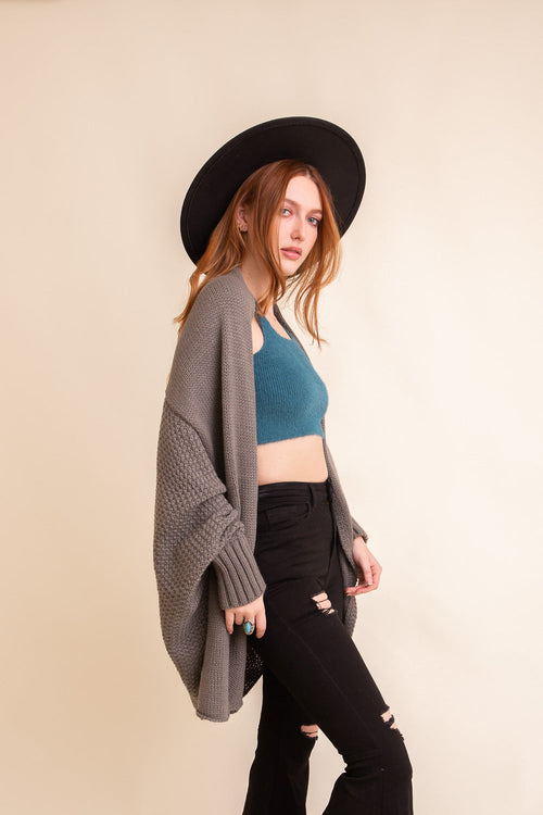 Woman wearing a black hat, gray cardigan, blue top, and black pants on a beige background