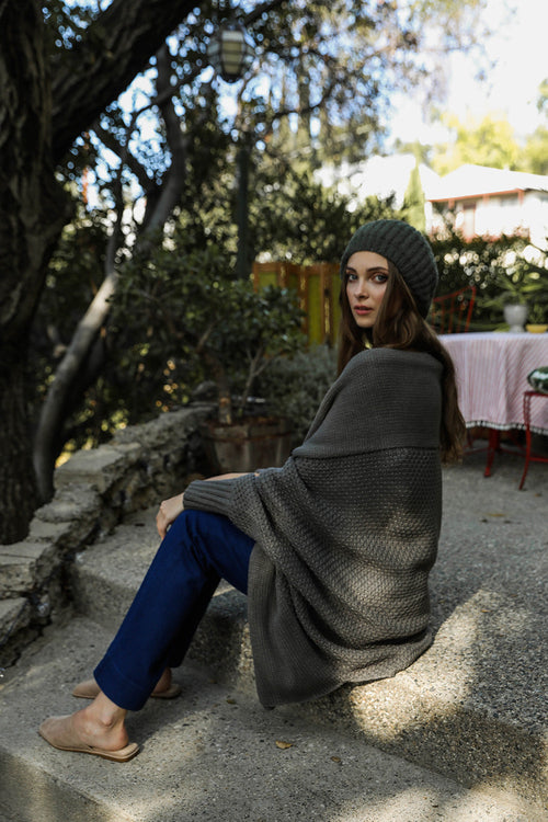 Woman sitting on steps outdoors wearing a gray poncho and knit hat.