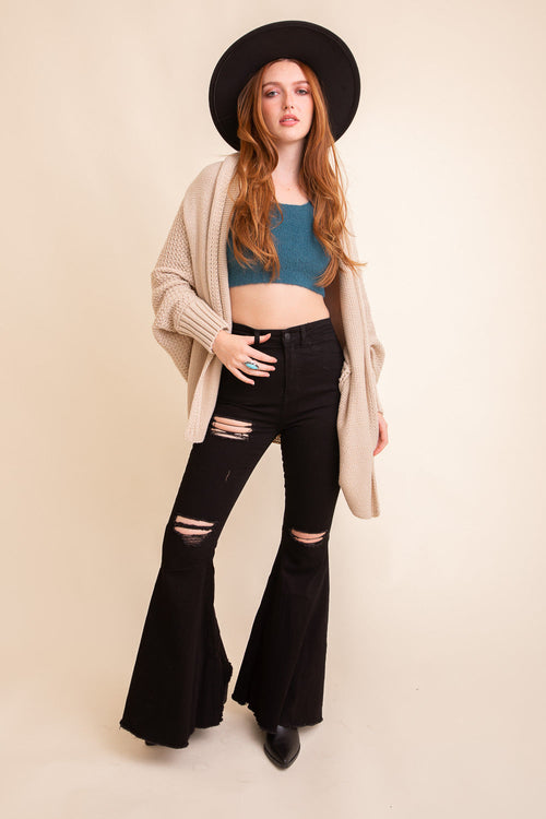 Woman wearing a black hat, beige cardigan, blue top, and black flared jeans on a beige background