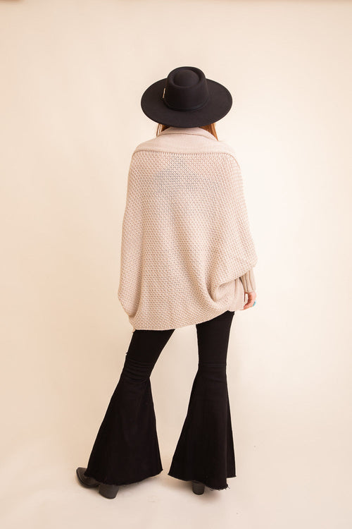 Person wearing a beige poncho and black wide-leg pants with a black hat on a beige background