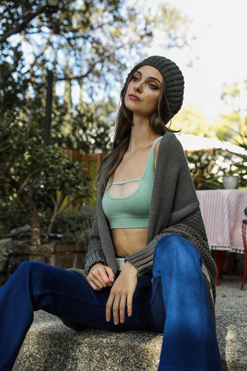 Woman sitting outdoors wearing a green sports bra and blue leggings with a gray cardigan.