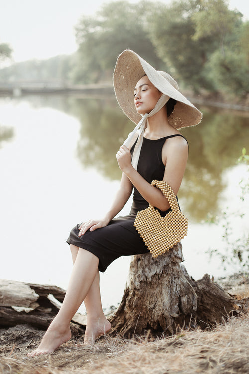 Woman sitting on a log by a river, wearing a wide-brimmed hat and holding a yellow handbag.
