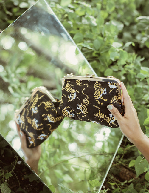 Person holding a black clutch with gold tiger pattern in front of a mirror against a green leafy background
