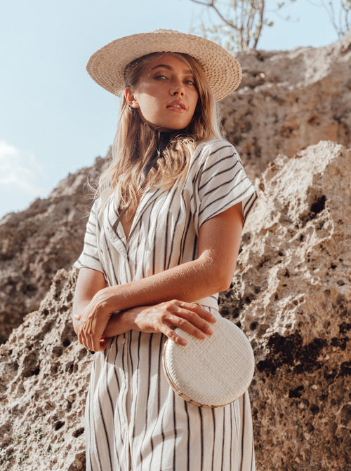 Woman in a striped dress and straw hat holding a circular bag against a rocky background