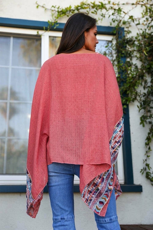 Woman wearing a pink poncho with patterned trim standing in front of a window.