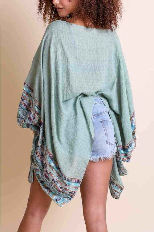 Woman wearing a light green poncho with patterned sleeves against a beige background