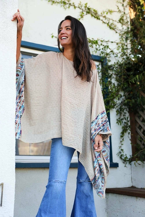 Woman wearing a beige poncho with floral sleeves in front of a white wall with greenery.