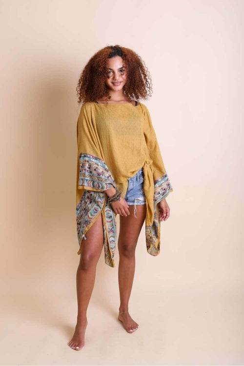 Woman wearing a mustard yellow top with floral sleeves and denim shorts on a beige background