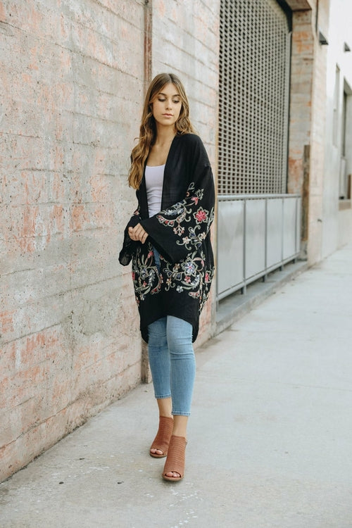 Woman wearing a black floral cardigan, white top, blue jeans, and brown sandals on a sidewalk.