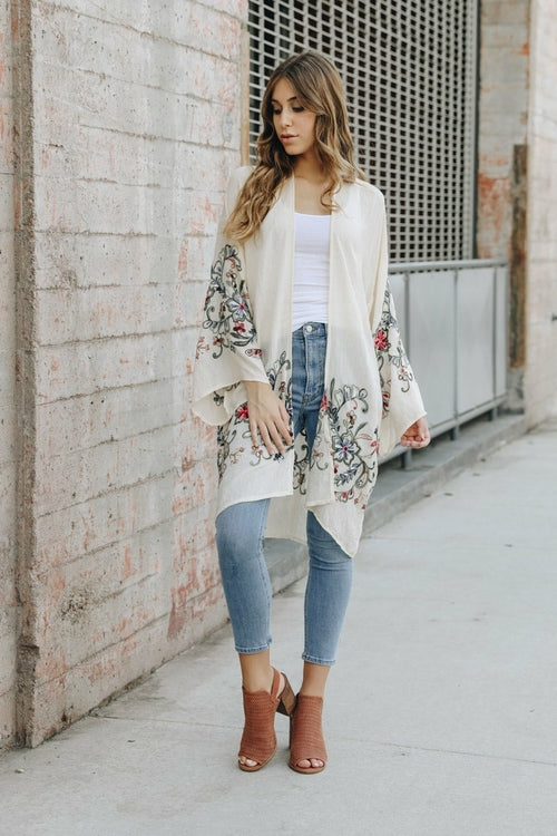Woman wearing a floral kimono over a white top and blue jeans, standing against a textured wall.