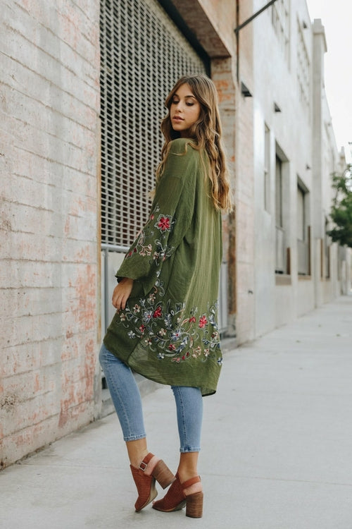 Woman wearing a green embroidered dress with blue jeans against a brick wall.