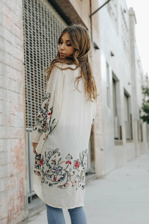 Woman wearing a long, light-colored coat with floral embroidery on a city street.