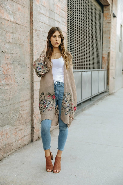Woman wearing a long embroidered cardigan, white top, blue jeans, and brown shoes on a sidewalk.